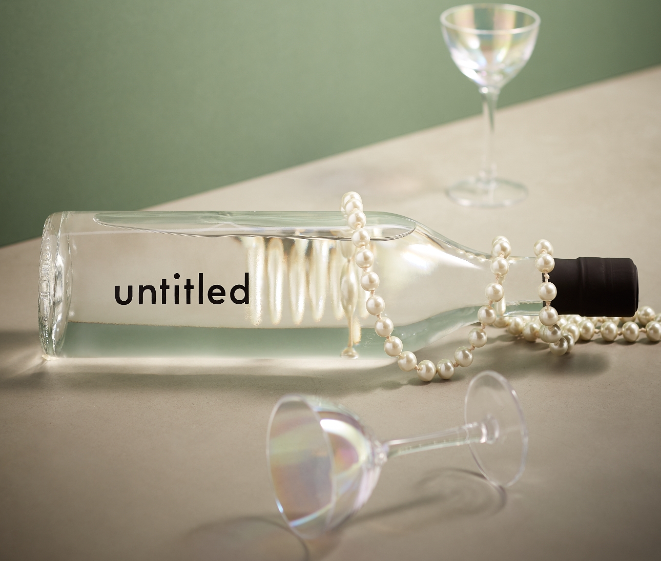 clear bottle of drink laying on it's side with two glasses next to it a a pearl necklace draped over it