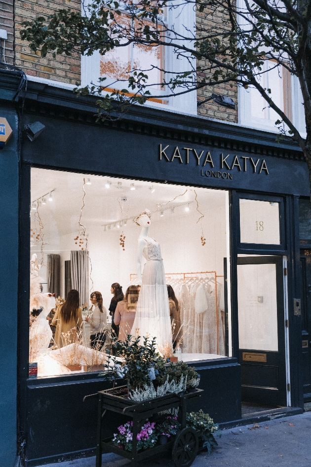 shop front of store with black wood trim, and models in the window and flowers in a wheelbarrow outiside