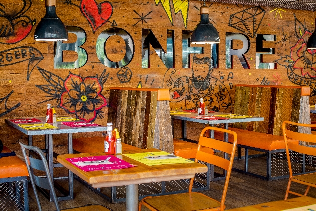 large wooden wall with tattoo print on it with wooden booths and colourful metal chairs