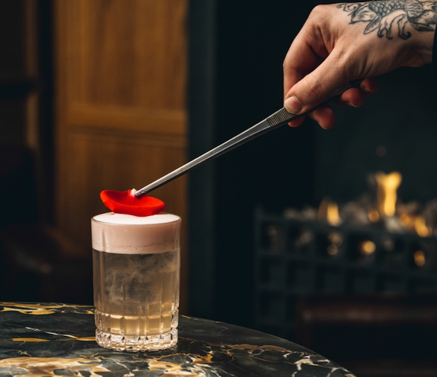 cocktail in a short glass frothy head and red rose petal being placed on top