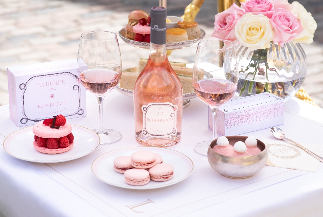 group image of a bottle of rose on a table with a plate of pink macarons and other desserts