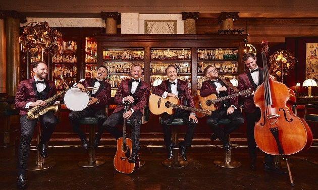 group of men suited in evening wear with guitars sat down on barstools in wood-panelled bar