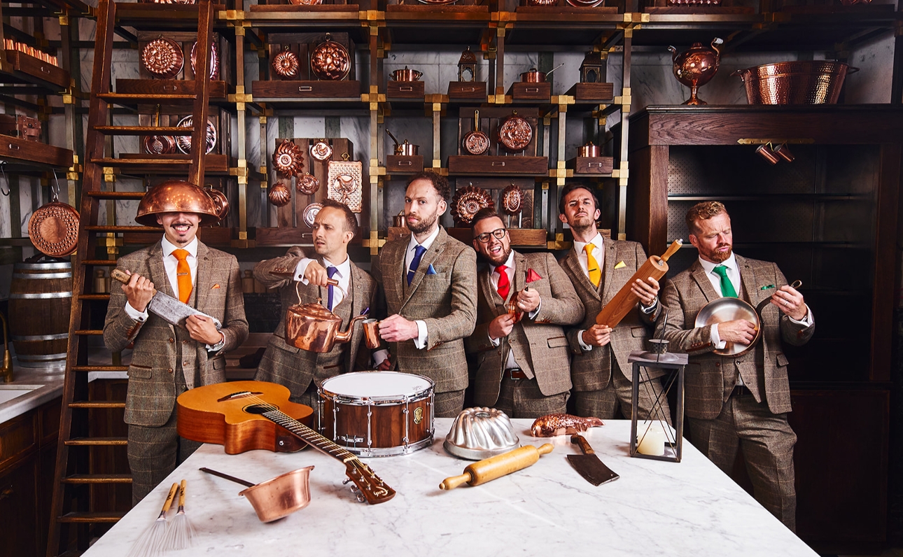 men in tweed suits in a cellar playing music on pots and pans 