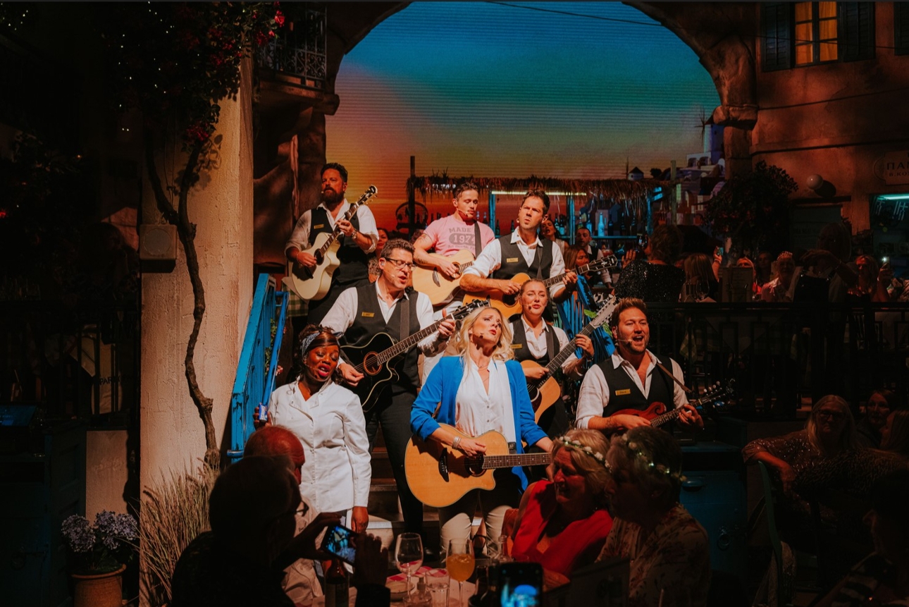 cast from Mamma Mia perfoming group song with guitars on set