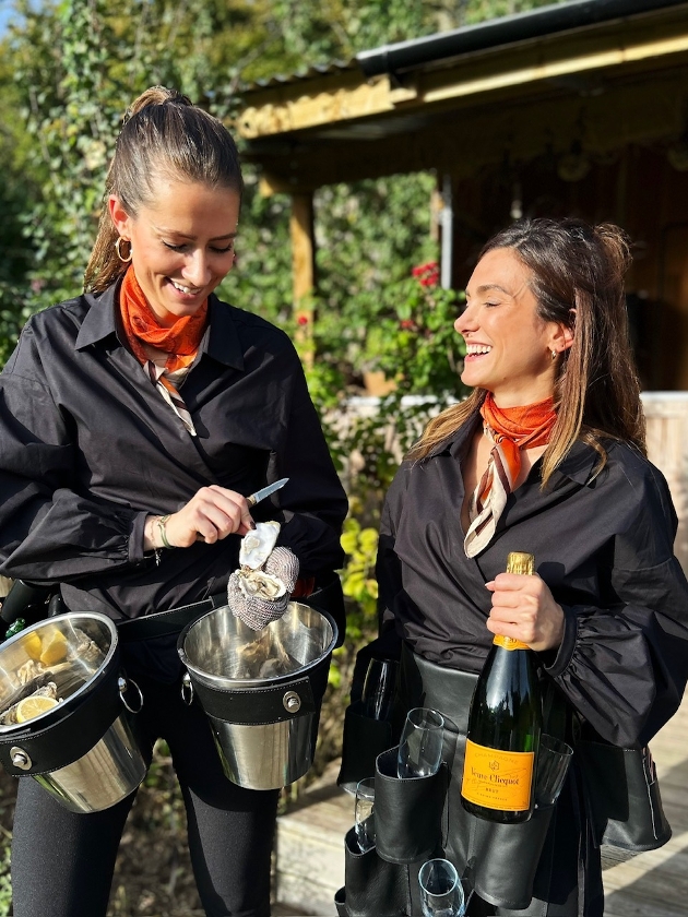 woman with oysters in a bucket,  next to woman with champagne and glasses