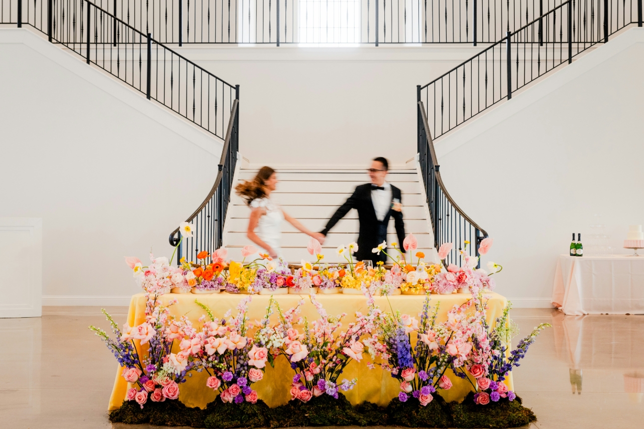 Couple running through a large hall behind a table set up with flowers